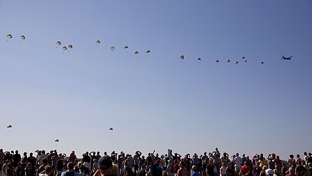 Publiek kijkt naar een rij parachutisten die zojuist uit een vliegtuig zijn gesprongen. 